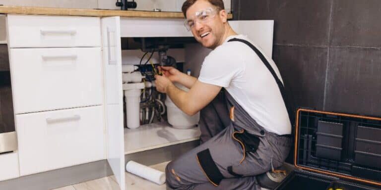 Professional plumber smiling while working on pipes under a kitchen sink