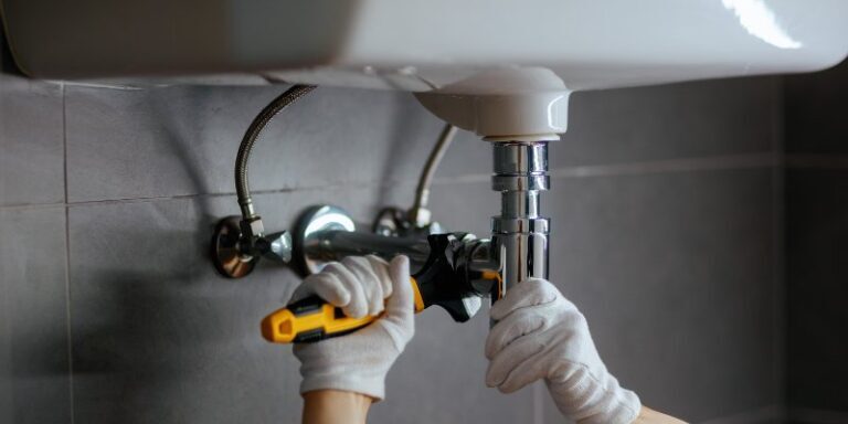 Plumber tightening a chrome drain pipe under a white sink with a wrench